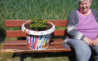 Seniorin sitzt auf einer Bank neben dem bemalten Erinnerungstopf