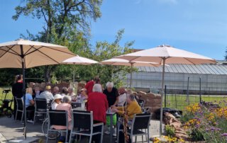 Zwei sommerliche Grillwochen mit fröhlicher Stimmung Gäste sitzen zwischen Blumenbeeten unter Sonnenschirmen